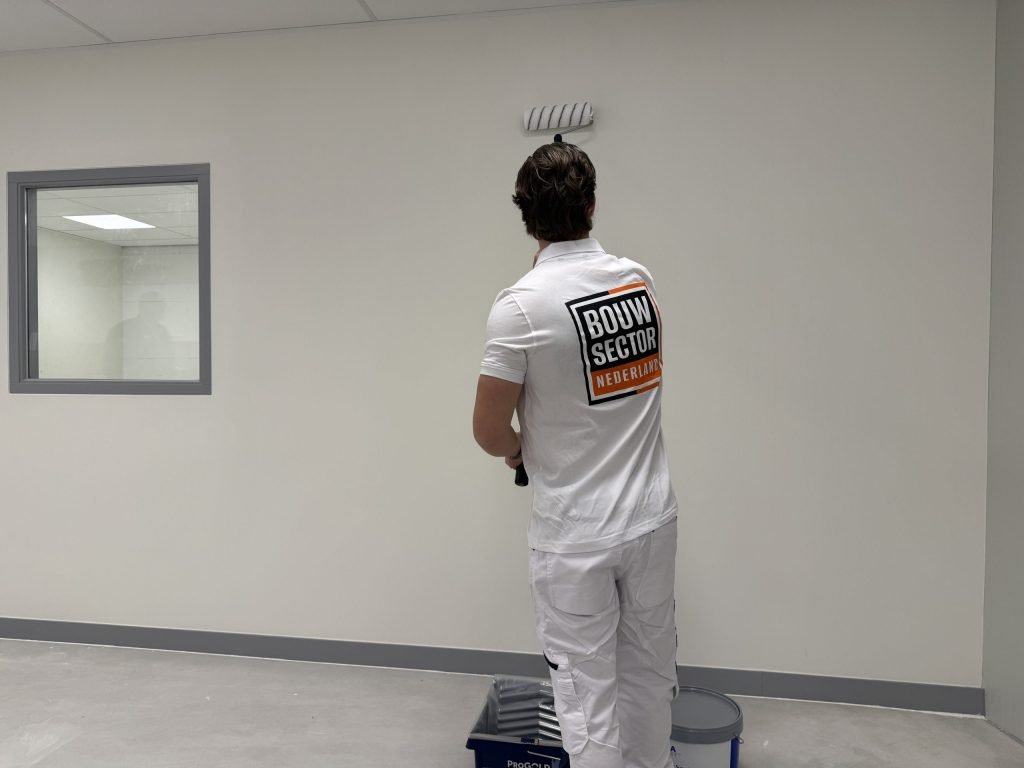 Schilder in witte werkkleding saust een lichte wand in een lege ruimte, met verfbak en emmer op de vloer