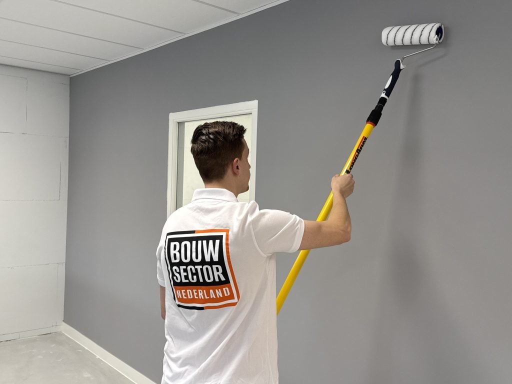 Schilder in een wit poloshirt van Bouwsector Nederland rolt grijze verf op een muur naast een deur