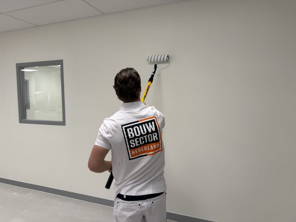 Schilder van Bouwsector Nederland rolt met een gele verlengsteel een lichte wand naast een binnenraam met grijs kozijn