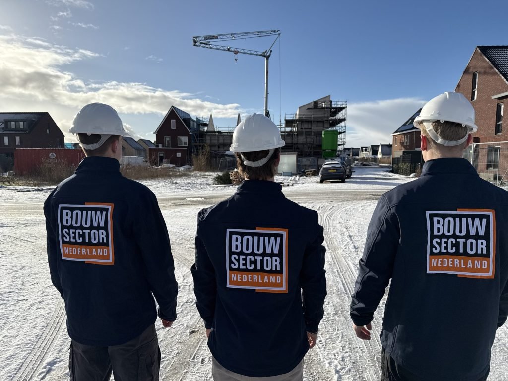Drie vakmensen van Bouwsector Nederland in donkerblauwe jas en witte helm kijken naar een nieuwbouwstraat met een kraan in de sneeuw