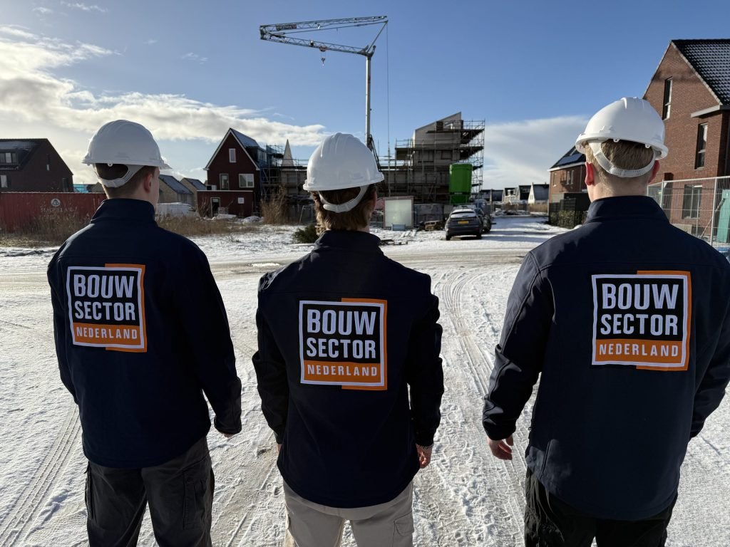 Drie vakmensen met Bouwsector Nederland op hun jas lopen over een besneeuwde bouwweg naar nieuwbouwwoningen